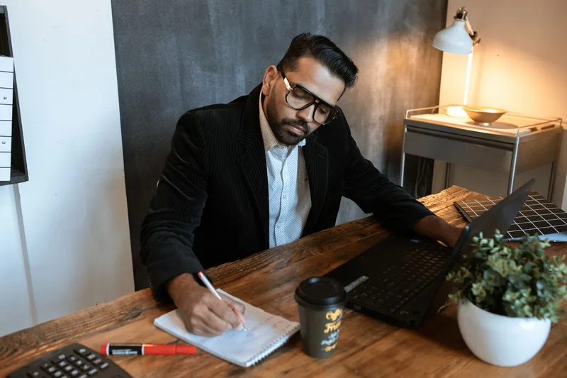A founder writing notes at a desk beside a laptop and a coffee cup, focused rather than inspired