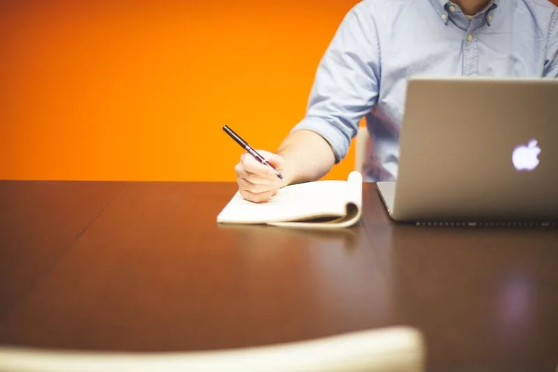 A person writing by hand in a notebook next to an open laptop, the kind of pause to weigh evidence before deciding whether it clears the threshold to start building