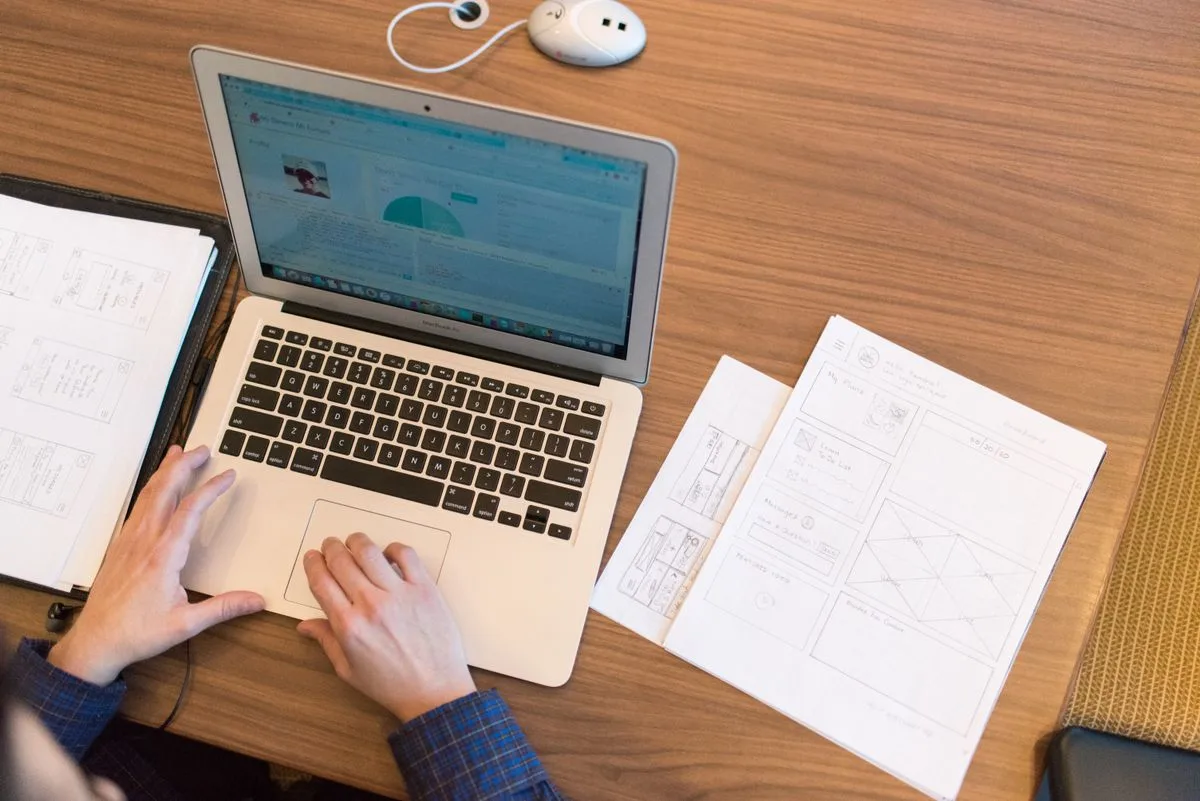 An overhead view of someone at a laptop showing a dashboard-style screen, with printed wireframe sketches spread on the desk beside it